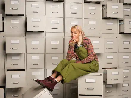 HD PC desktop wallpaper for the TV show High Potential: a young woman in green overalls sits on a filing cabinet amid rows of drawers, smiling thoughtfully.