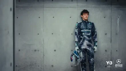 F1 driver in Mercedes-AMG race suit holding a helmet, leaning against a concrete wall — 2K Quad HD PC desktop wallpaper background