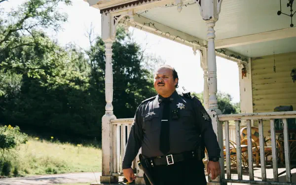 4K Ultra HD PC desktop wallpaper and background from the movie Bugonia: a uniformed police officer stands on a sunlit rural porch beside a wooden railing and trees.