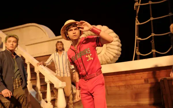 One Piece (Live Action) 2K Quad HD wallpaper: straw-hat captain in a red tracksuit scans the horizon aboard a wooden ship with two crewmates under warm stage lighting.