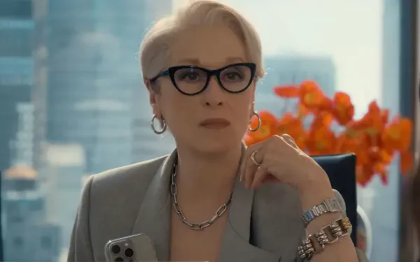 HD PC desktop wallpaper of a poised, silver‑haired actress in glasses and a gray suit from the movie The Devil Wears Prada 2, seated in a city office with orange flowers behind her.
