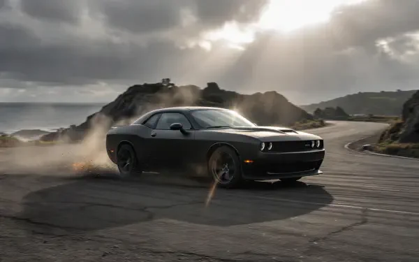 Black Dodge Challenger drifting on a coastal road, kicking up smoke beneath dramatic clouds — 2K Quad HD PC desktop wallpaper/background.