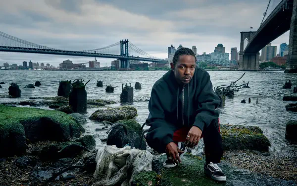 4K Ultra HD PC wallpaper: a hip-hop artist crouches on a rocky East River shore beneath the Manhattan Bridge in New York, moody urban skyline and textured waterfront background.