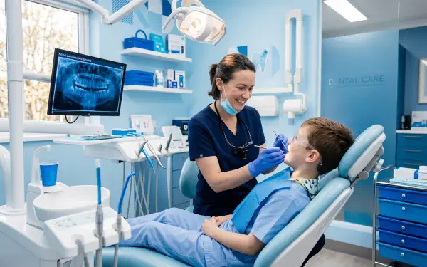 5K Ultra HD PC desktop wallpaper/background: dentist smiling while treating a young patient in a modern dental clinic, bright blue treatment room with an x-ray monitor.