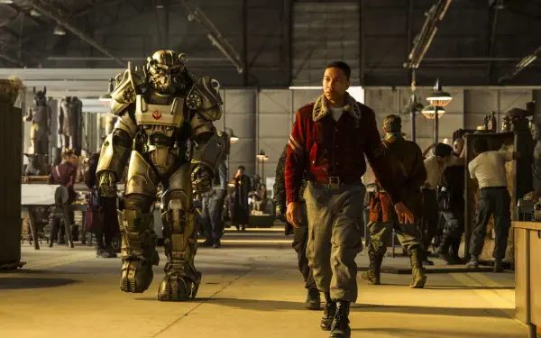 Man in a red coat strides through an industrial hangar beside a towering power armor suit, figures and gear in the background — Fallout (TV Show) HD PC desktop wallpaper and background.