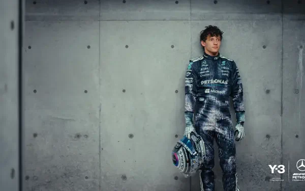 F1 driver in Mercedes-AMG race suit holding a helmet, leaning against a concrete wall — 2K Quad HD PC desktop wallpaper background