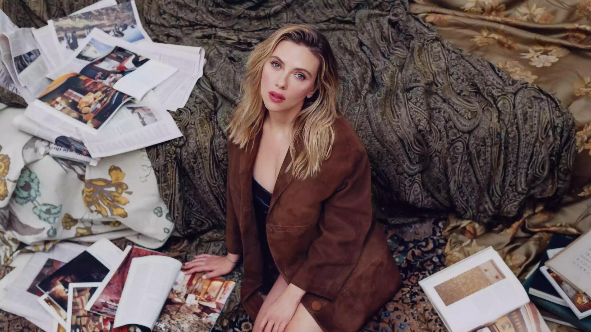 Portrait of a blonde actress and celebrity seated among scattered photographs and draped fabrics, gazing at the camera — 4K Ultra HD PC desktop wallpaper.