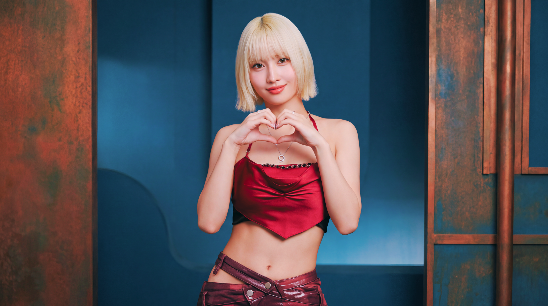 5K Ultra HD desktop wallpaper: blonde woman with a short bob in a red satin crop top and low-rise pants, making a heart with her hands—K-pop–inspired styling against a blue-copper studio backdrop.