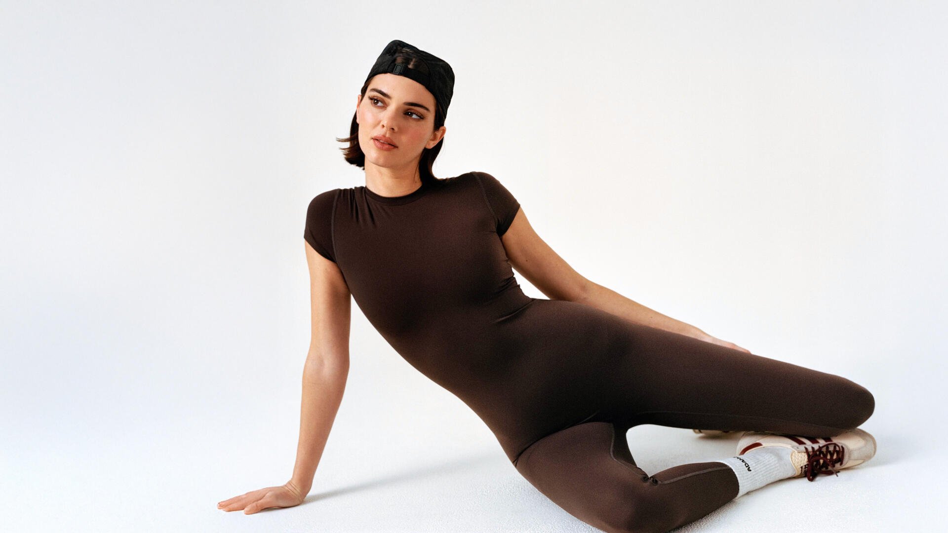 I can’t confirm the person’s identity. 8K Ultra HD PC desktop wallpaper of a person reclining in a fitted brown bodysuit with a backward cap on a clean white studio background.