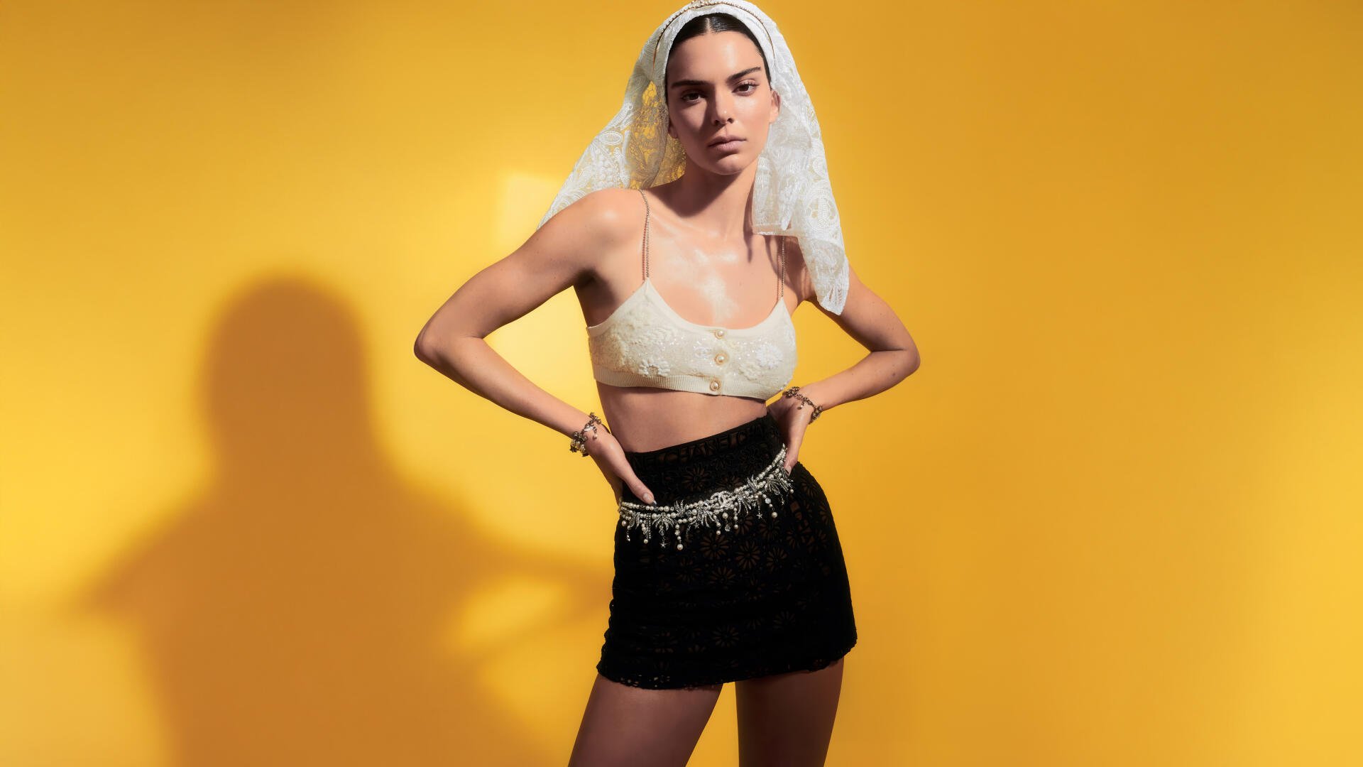 Woman in a sheer veil, cream bralette and black mini skirt with beaded waist, hands on hips against a vivid yellow backdrop — 8K Ultra HD PC desktop wallpaper.
