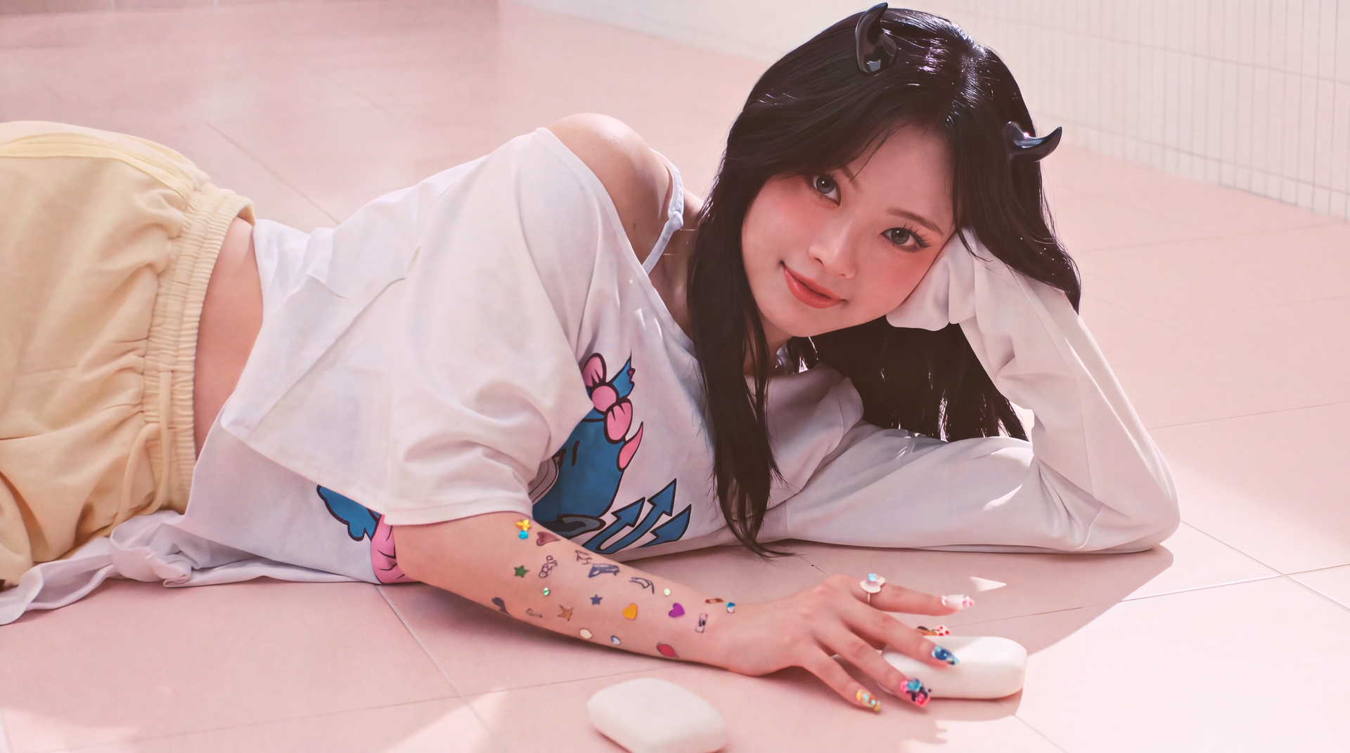 5K desktop wallpaper of a smiling young woman with long dark hair reclining on a pale pink floor, white tee and pastel shorts, colorful stickers on her arm — cute K-pop–inspired aesthetic.
