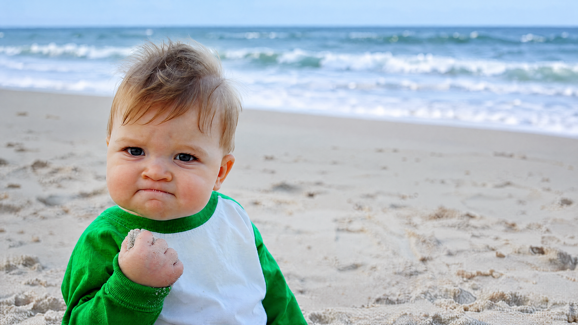 Fist-pumping baby meme with a triumphant expression on a sandy beach, classic success-meme vibe; 4K Ultra HD PC desktop wallpaper/background.