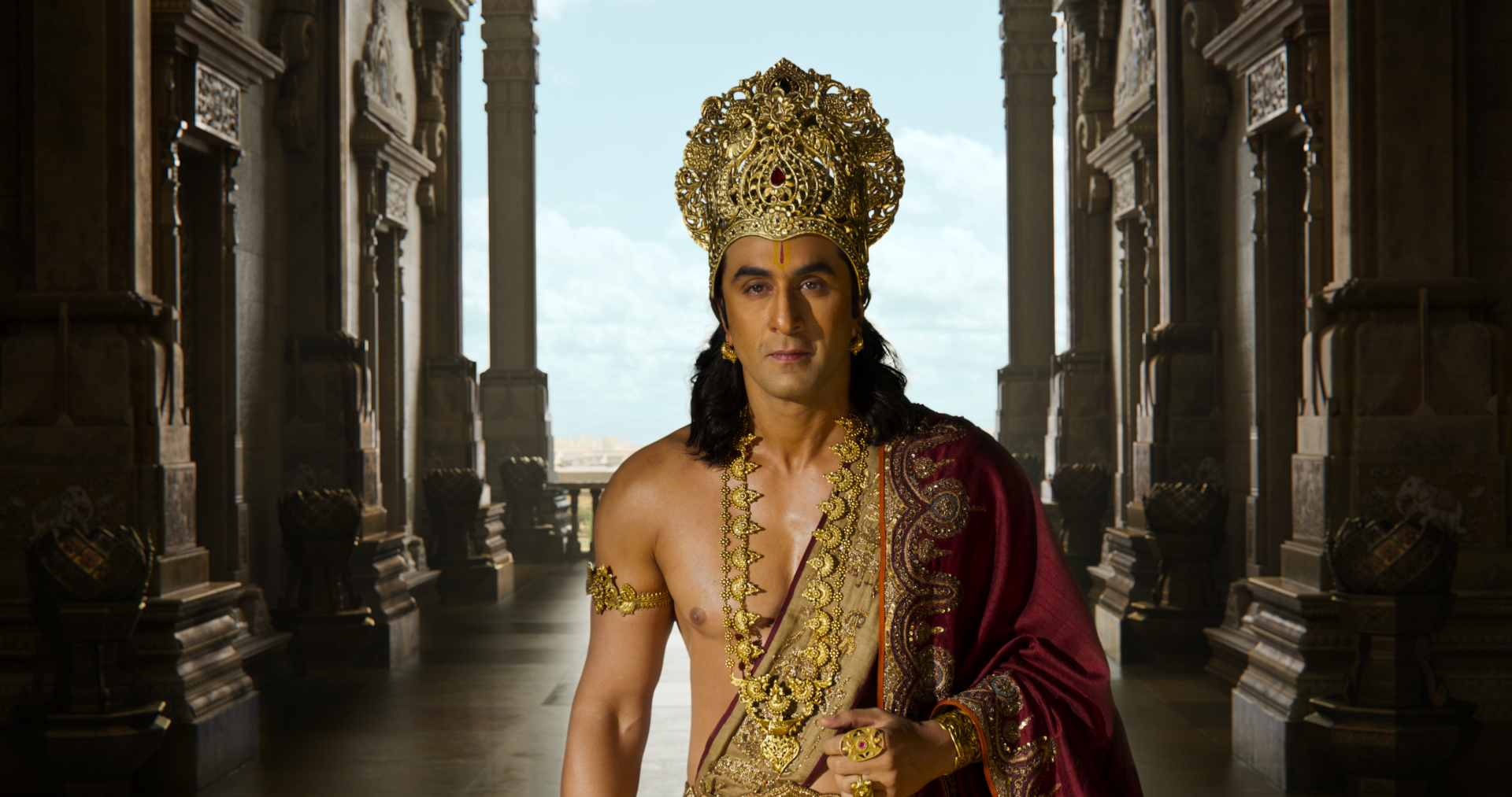 Actor in Ramayana (2026) as a royal king wearing an ornate gold crown and red cloak in a palatial corridor — 4K Ultra HD PC desktop wallpaper background.