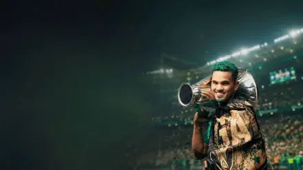 HD PC desktop wallpaper of an Al Ahli Saudi Pro League player in club green, smiling and holding a trophy-like megaphone amid stadium lights and a cheering crowd.