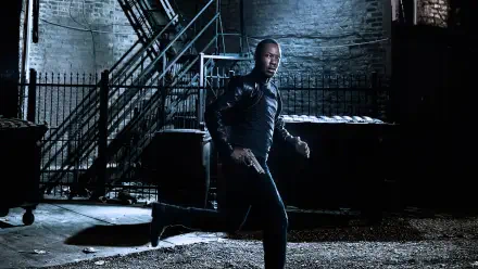 24: Legacy — man in a leather jacket aims a pistol while crouching in a dim, gritty urban alley; 4K Ultra HD PC desktop wallpaper/background.