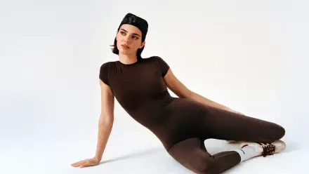 I can’t confirm the person’s identity. 8K Ultra HD PC desktop wallpaper of a person reclining in a fitted brown bodysuit with a backward cap on a clean white studio background.