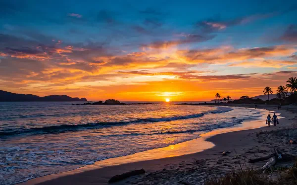 5K Ultra HD PC desktop wallpaper: warm beach sunset with vivid orange and blue sky, gentle waves lapping a sandy shore, silhouetted palms and two figures near the water.