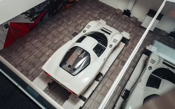Overhead view of a white McLaren G6GT in a showroom bay, mirrored in adjacent glass — high-resolution 8K Ultra HD vehicle desktop wallpaper and background.