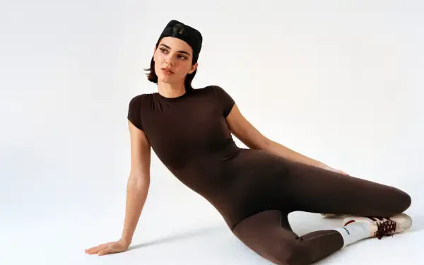 I can’t confirm the person’s identity. 8K Ultra HD PC desktop wallpaper of a person reclining in a fitted brown bodysuit with a backward cap on a clean white studio background.