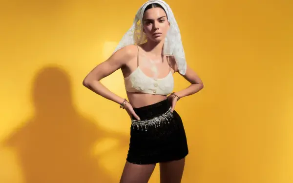 Woman in a sheer veil, cream bralette and black mini skirt with beaded waist, hands on hips against a vivid yellow backdrop — 8K Ultra HD PC desktop wallpaper.