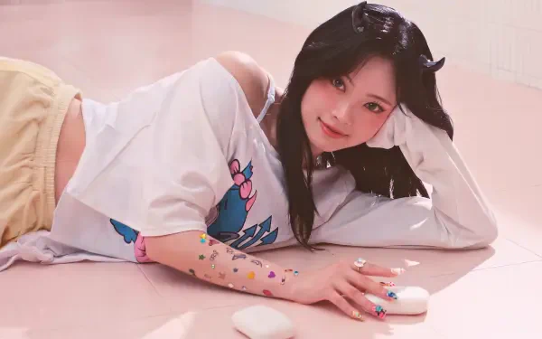 5K desktop wallpaper of a smiling young woman with long dark hair reclining on a pale pink floor, white tee and pastel shorts, colorful stickers on her arm — cute K-pop–inspired aesthetic.