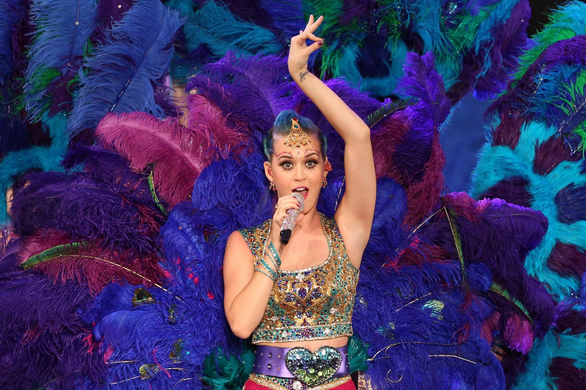 Vibrant HD wallpaper featuring Katy Perry performing, adorned in a dazzling outfit with colorful feathers behind her, capturing the energy of her music and stage presence.