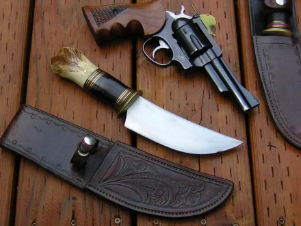 A close-up of a knife with a decorative handle rests on a wooden surface alongside a revolver and a leather sheath, creating a striking man-made composition.