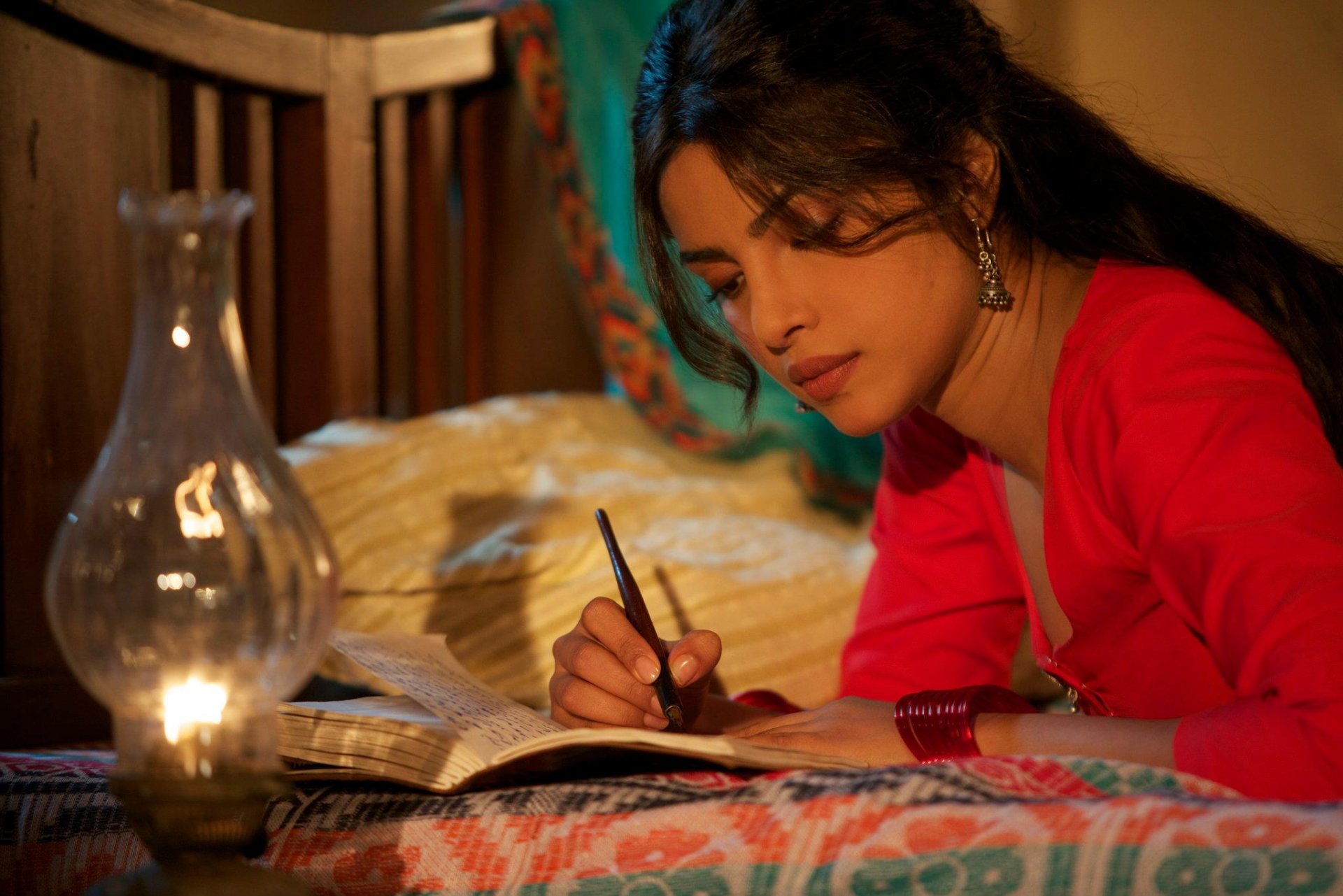 A captivating HD wallpaper featuring Priyanka Chopra, elegantly dressed in red, writing in a notebook by lantern light, set against a cozy, warmly lit backdrop.