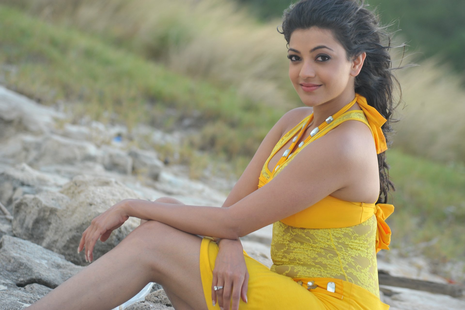 HD desktop wallpaper and background featuring celebrity Kajal Aggarwal sitting outdoors on rocky terrain, wearing a yellow dress, and smiling at the camera.