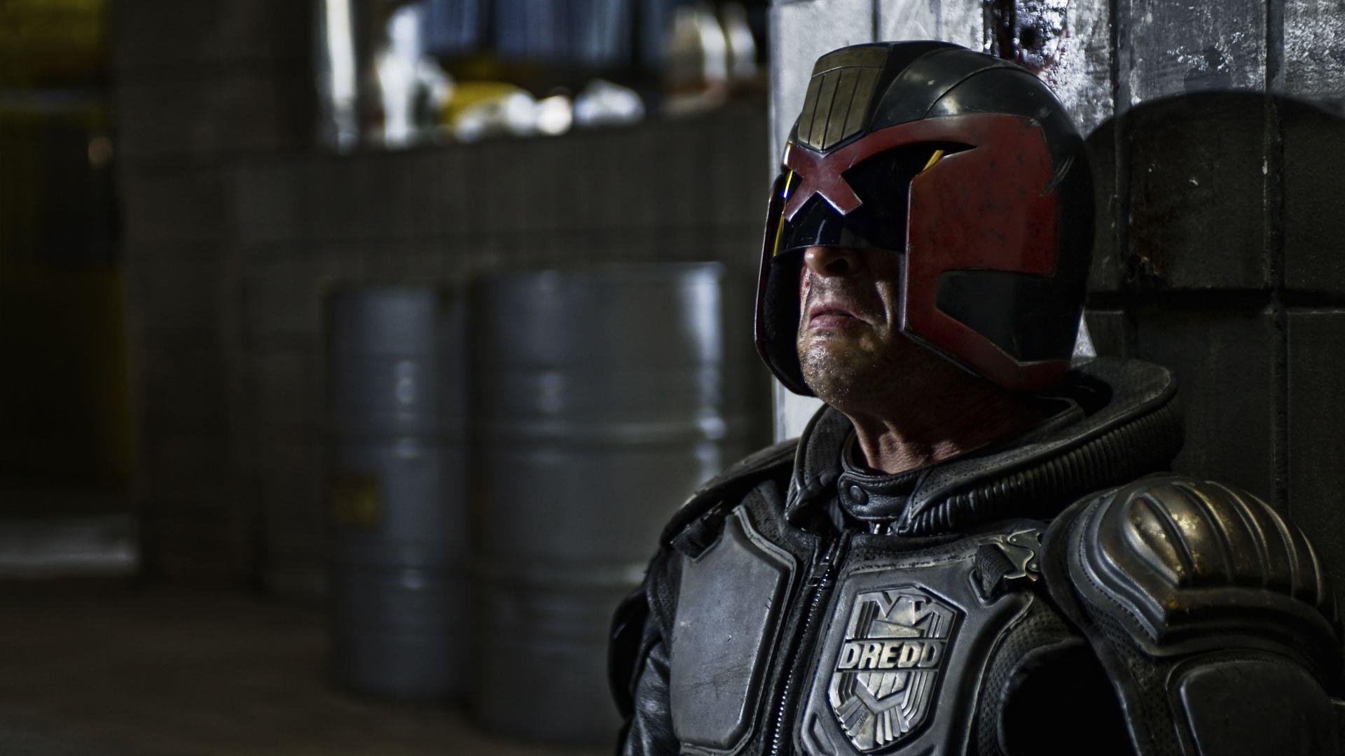 HD PC desktop wallpaper from the movie Dredd: close-up of armored Judge Dredd in helmet and visor, standing in a dim industrial corridor with stacked barrels.