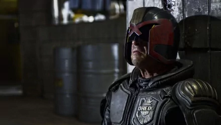 HD PC desktop wallpaper from the movie Dredd: close-up of armored Judge Dredd in helmet and visor, standing in a dim industrial corridor with stacked barrels.