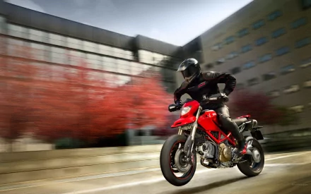 HD PC desktop wallpaper of a red motorcycle vehicle speeding down a blurred urban street, rider in black gear with autumn trees and modern buildings in background.