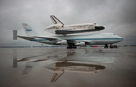 A NASA aircraft transports the Space Shuttle Discovery, reflected on a wet runway under a cloudy sky. This stunning image serves as a 4K Ultra HD desktop wallpaper.