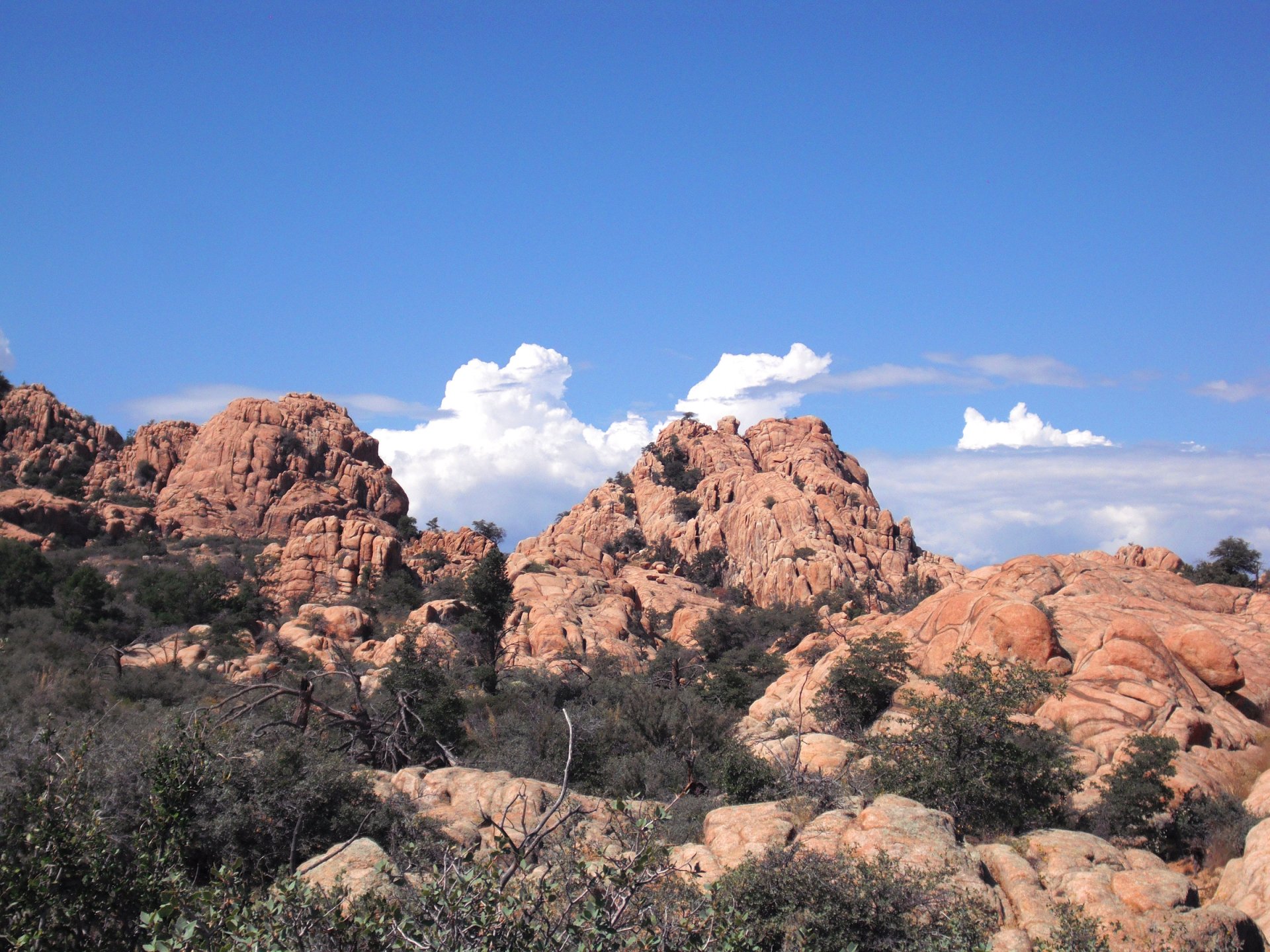HD PC desktop wallpaper background: sunlit red‑rock mountain landscape with sparse shrubs beneath a bright blue sky dotted with white clouds.