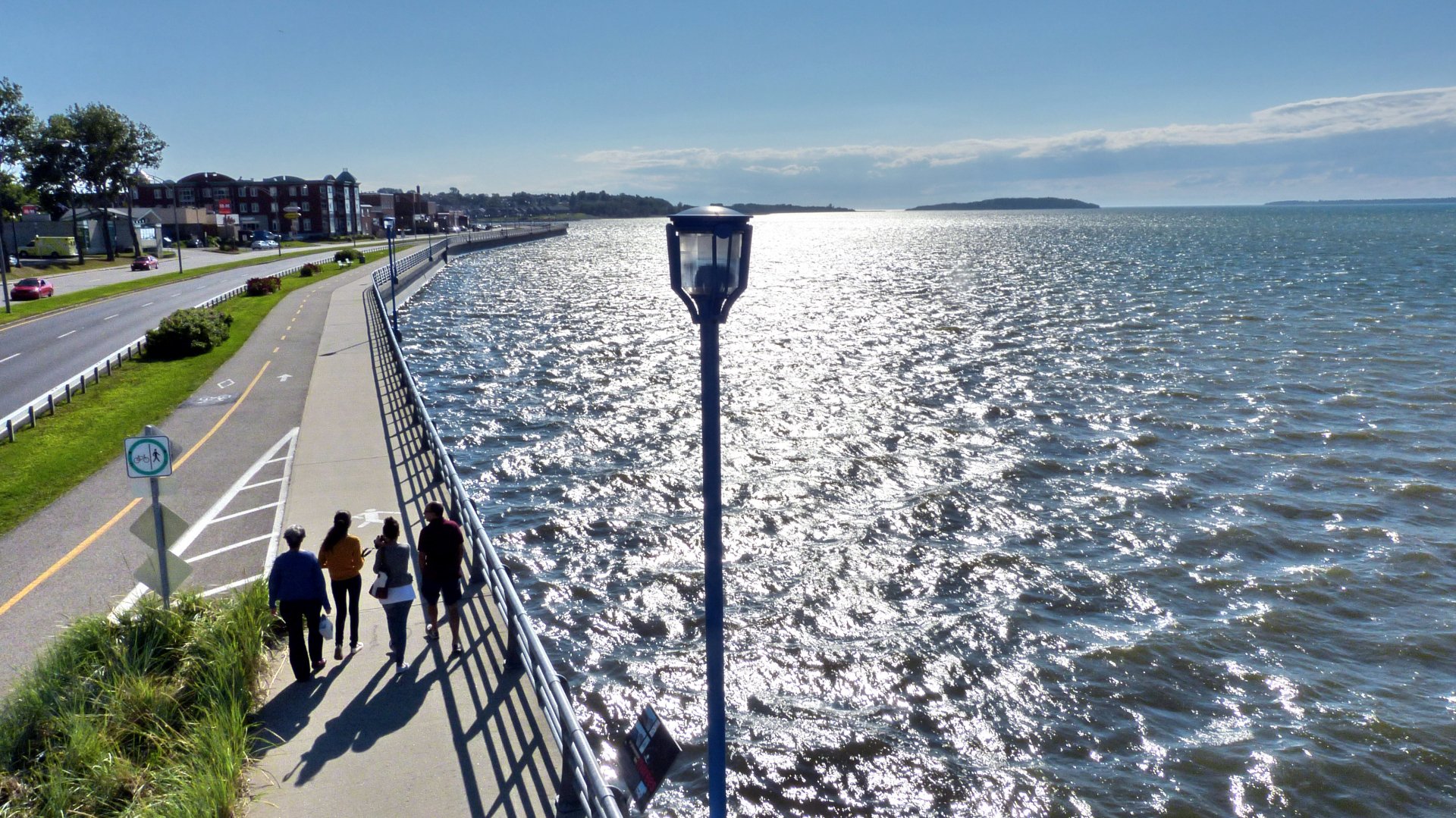 Place photography of a coastal promenade at midday, people walking beside a sun-sparkled sea, lamppost and distant town; 4K Ultra HD PC Desktop Wallpaper and Background.