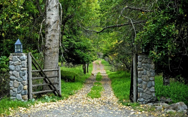 HD desktop wallpaper showing a nature path lined with trees, framed by a stone gate, leading down a quiet road through lush greenery.