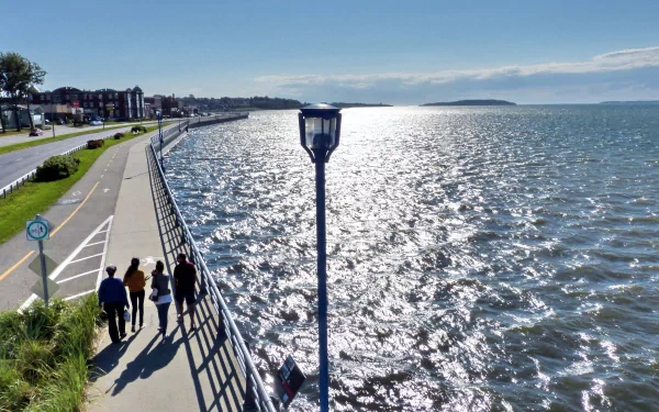 Place photography of a coastal promenade at midday, people walking beside a sun-sparkled sea, lamppost and distant town; 4K Ultra HD PC Desktop Wallpaper and Background.