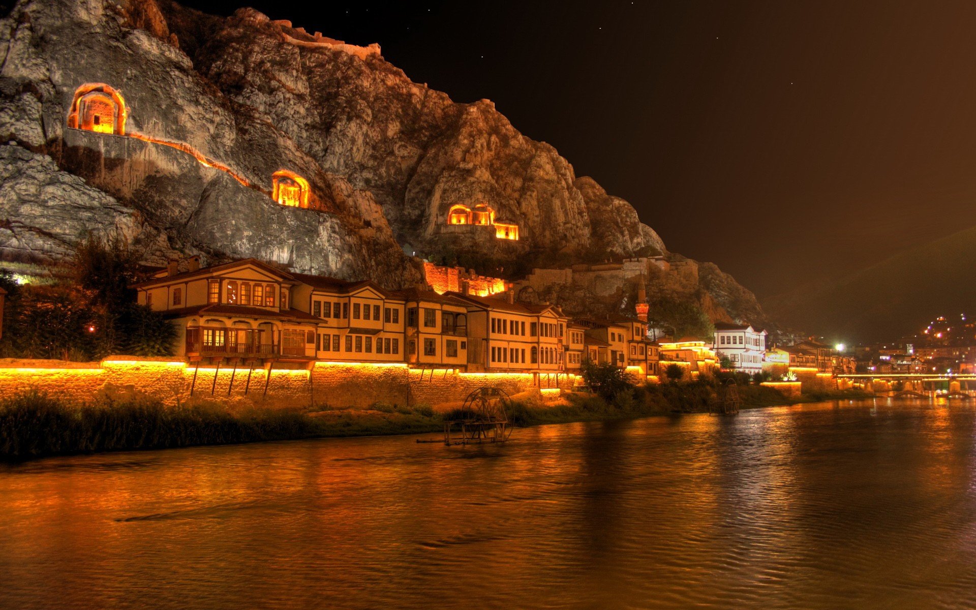 HD PC desktop wallpaper showing a man-made town illuminated at night, nestled against rocky cliffs along a river.