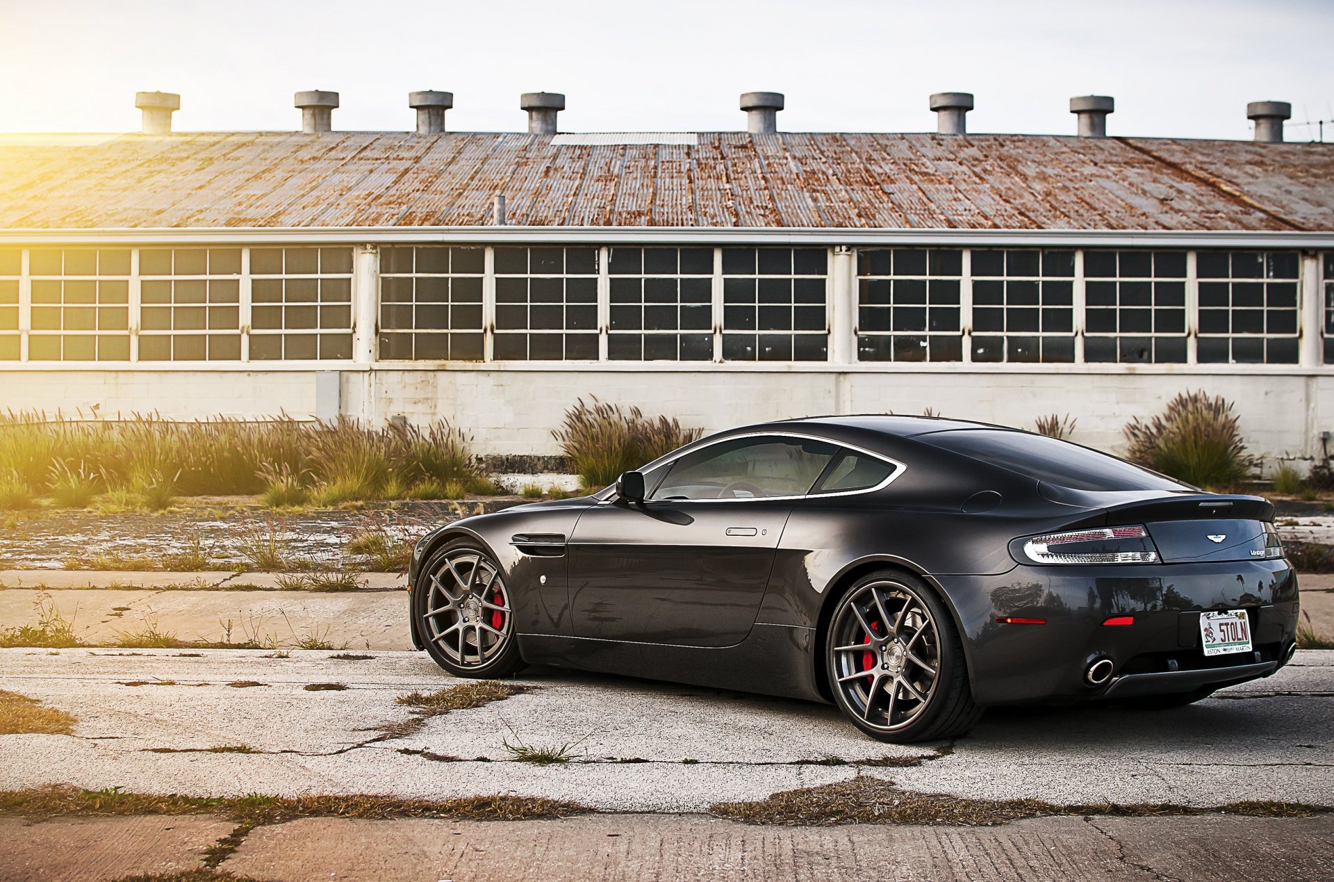 A sleek black Aston Martin V8 Vantage parked in front of an industrial building, captured in an HD PC desktop wallpaper and background.