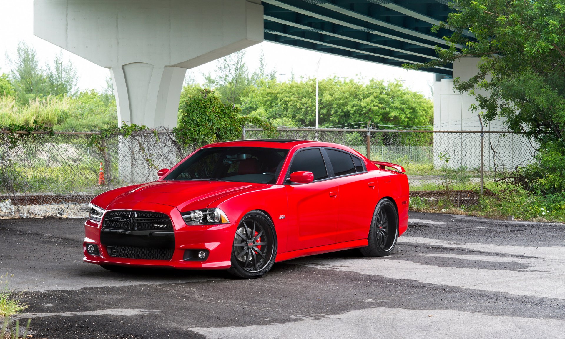 Red Dodge Charger parked beneath an overpass, wet pavement and chain-link fence in the background — high-detail 4K Ultra HD PC desktop wallpaper.