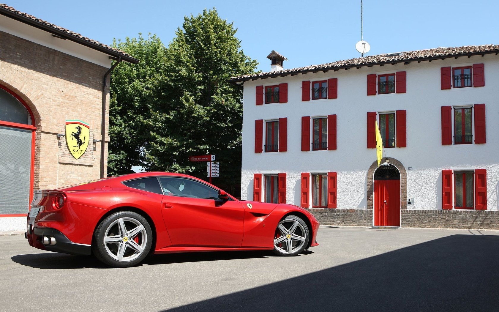 Red Ferrari F12berlinetta parked in a sunlit courtyard in front of a white building with red shutters — HD PC desktop wallpaper background.