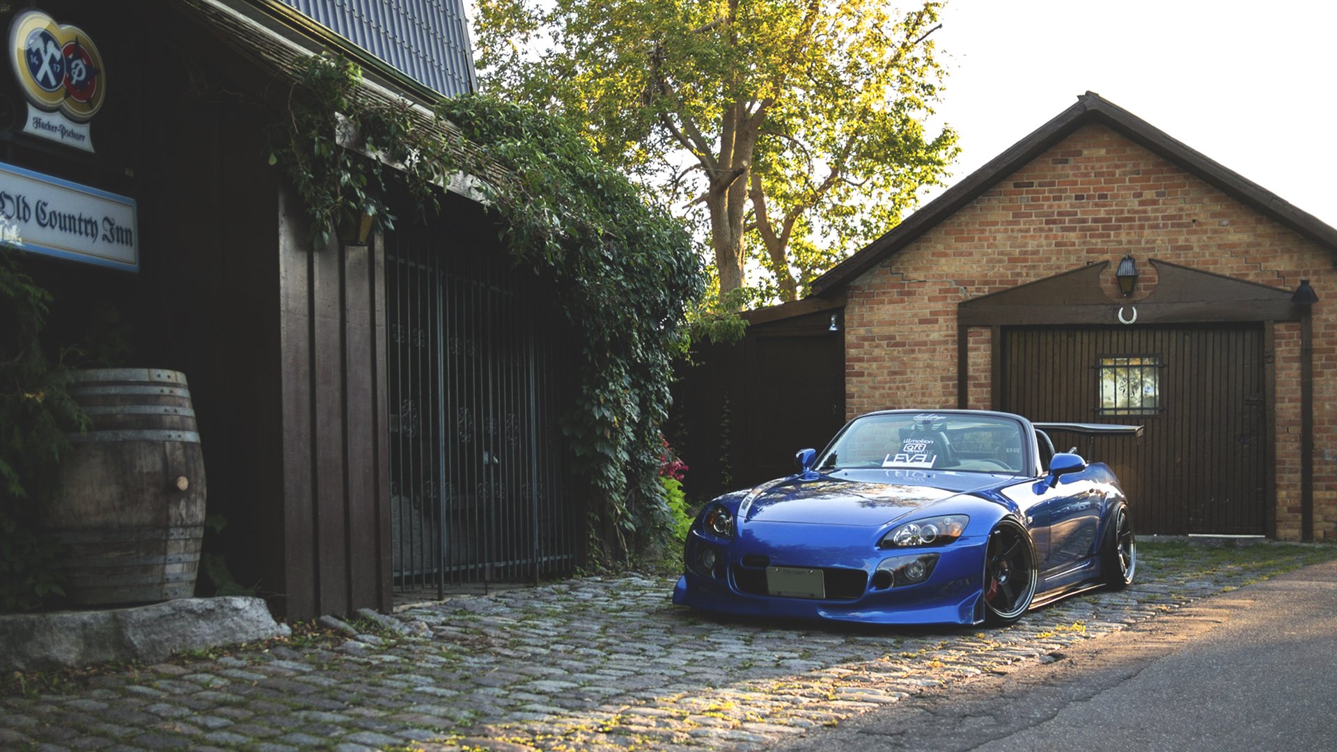 HD PC desktop wallpaper featuring a blue Honda sports car parked on a cobblestone driveway beside rustic buildings and greenery.