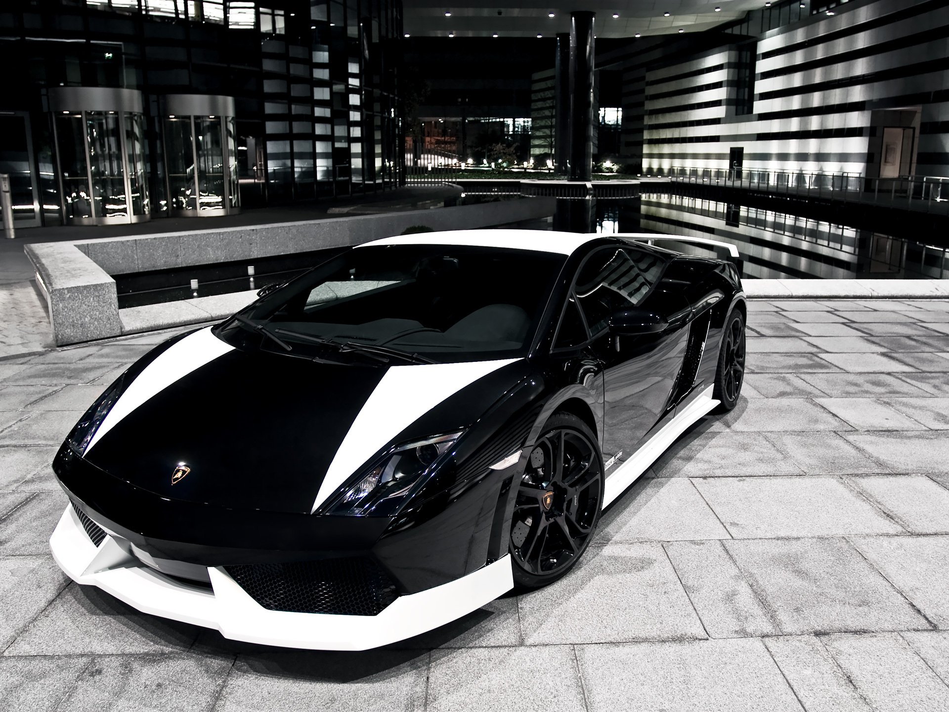 HD PC desktop wallpaper/background showing a black-and-white Lamborghini Gallardo vehicle parked on a tiled urban plaza at night with reflective modern buildings.