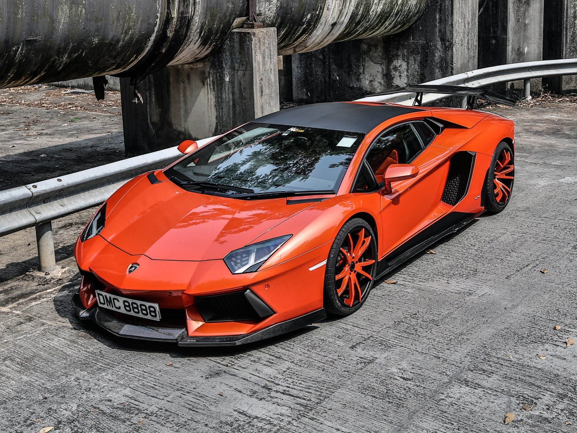 HD PC desktop wallpaper featuring a vibrant orange Lamborghini Aventador parked on an urban street under industrial pipes.