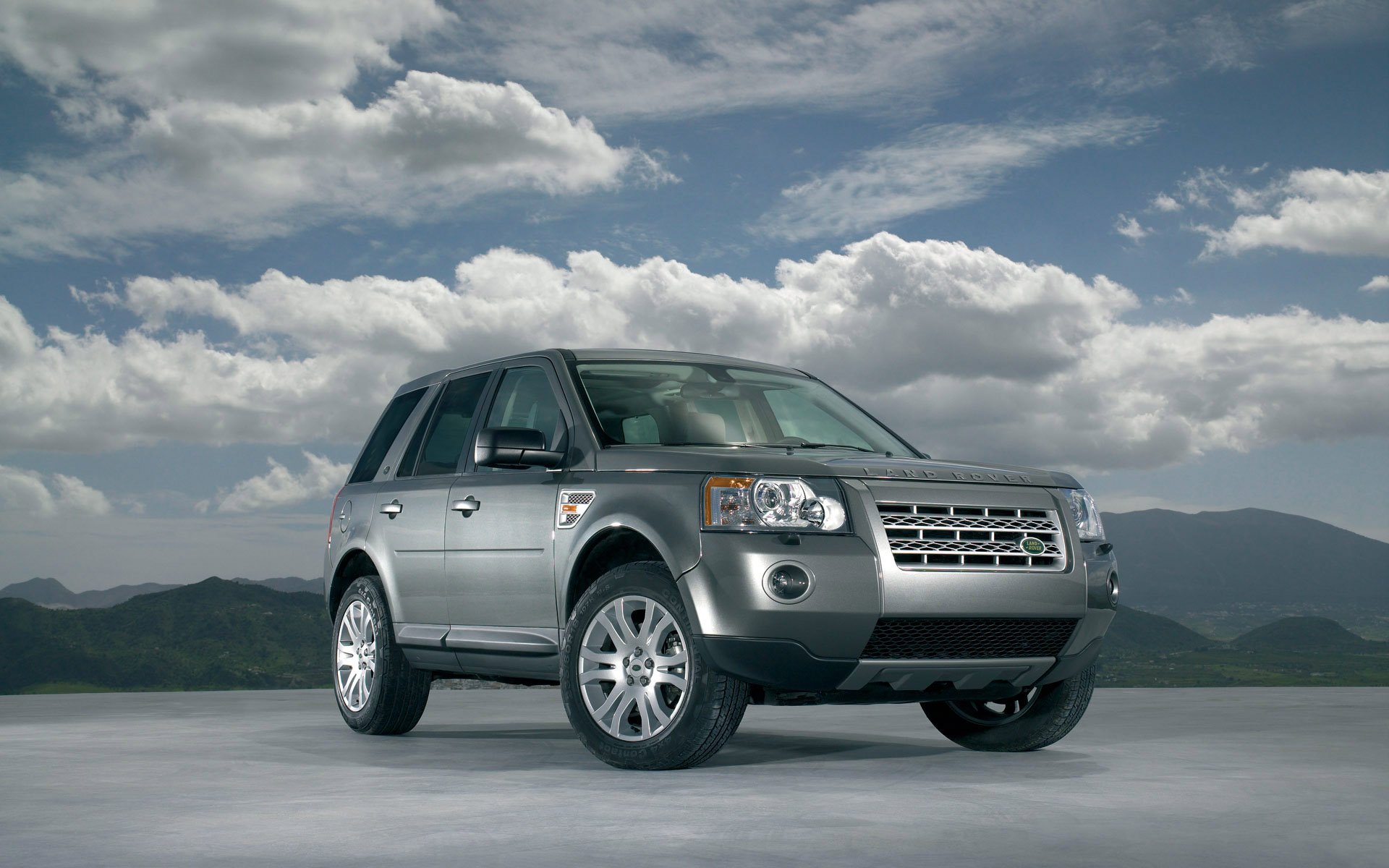Silver Land Rover vehicle parked on flat pavement beneath dramatic clouds and distant hills — HD PC desktop wallpaper/background.