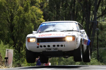 HD desktop wallpaper featuring a Ford Capri vehicle captured mid-air during a rally event, surrounded by lush green trees.