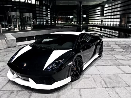 HD PC desktop wallpaper/background showing a black-and-white Lamborghini Gallardo vehicle parked on a tiled urban plaza at night with reflective modern buildings.