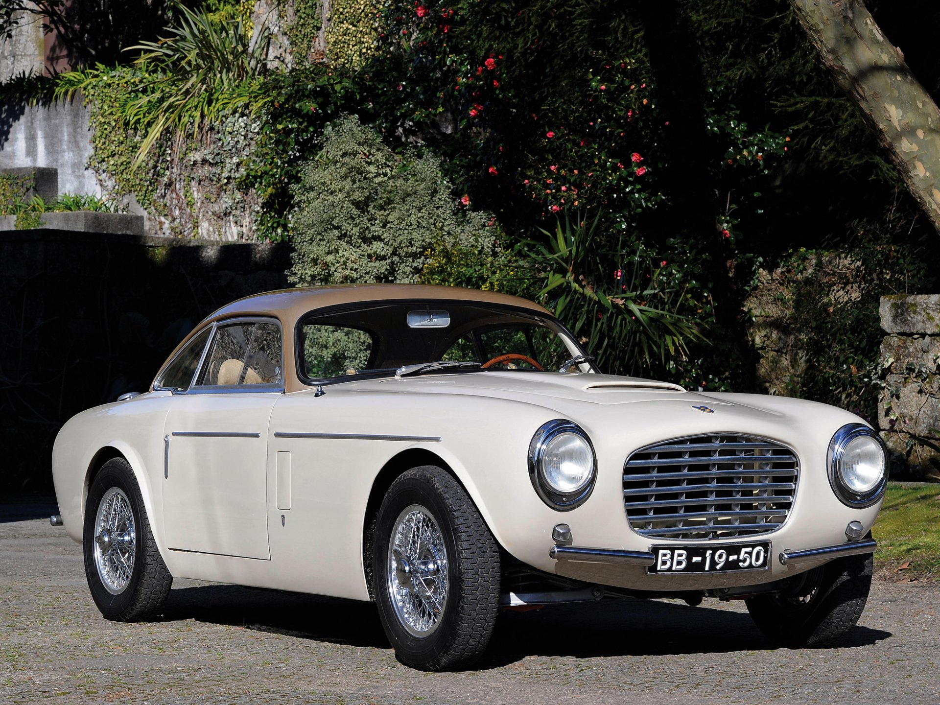 HD PC desktop wallpaper of a white Siata vehicle parked by a stone wall and lush garden, front three-quarter view showcasing chrome grille, round headlights and wire-spoke wheels.