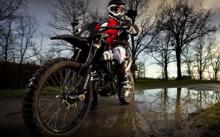 A motocross rider in full gear atop a dirt bike, standing on a wet path with bare trees in the background. HD desktop wallpaper and background.