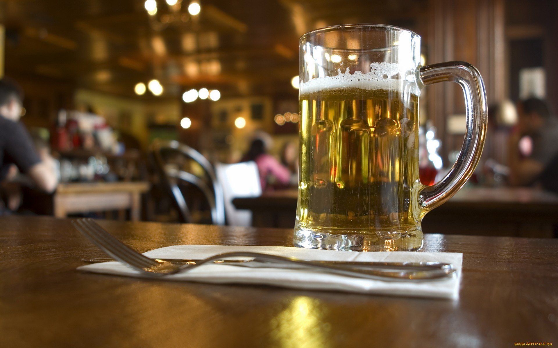 HD PC desktop wallpaper showcasing a clear glass mug of beer on a wooden table with a fork and knife, set against a warmly lit, blurred pub background.